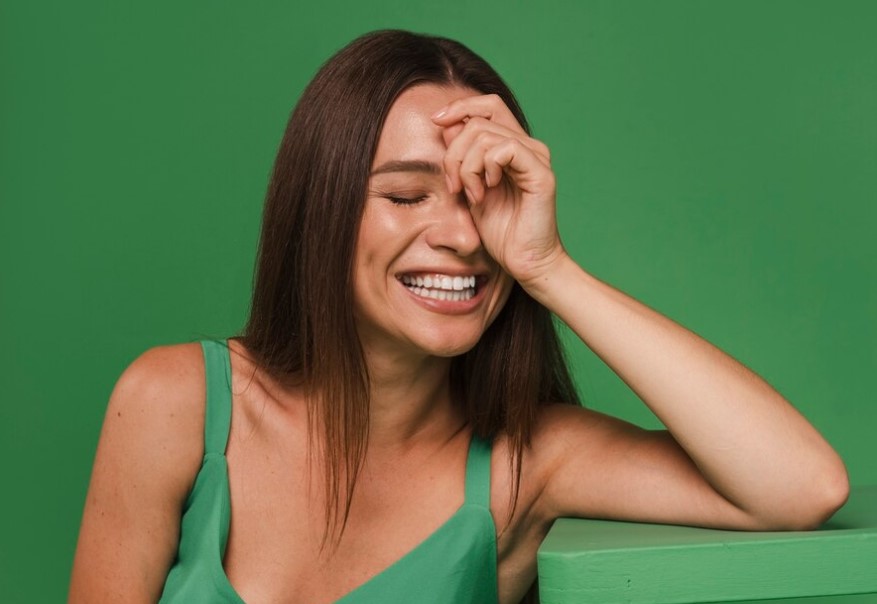 Mujer con fondo verde y vestido verde se rie mientras se tapa parte de la cara