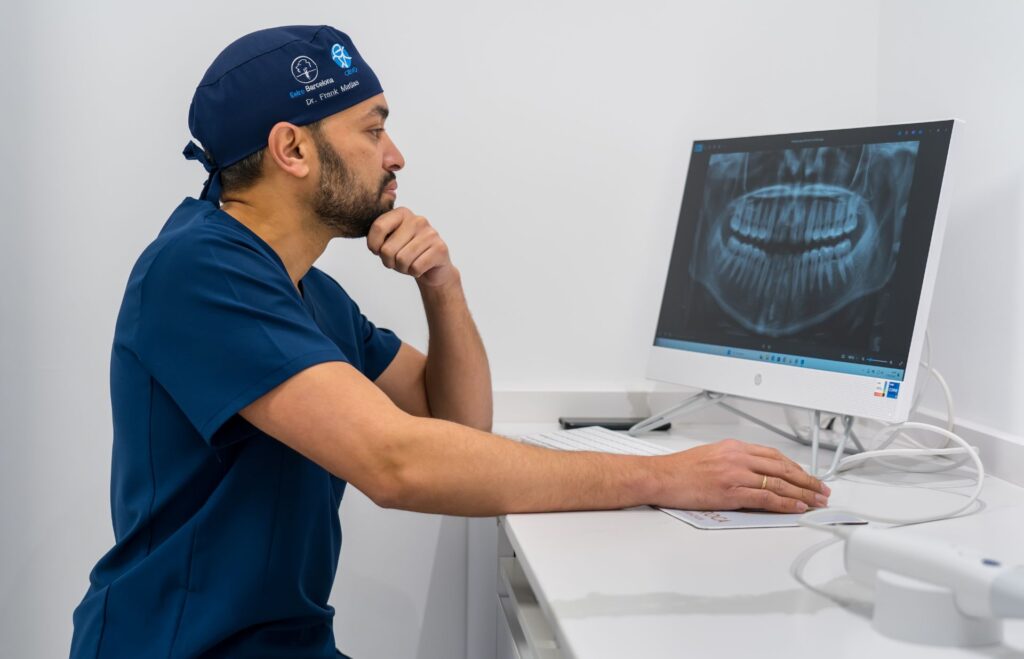 Dentista en camisa azul revisando una radiografía de dientes en su computadora mientras está sentado en un escritorio.
